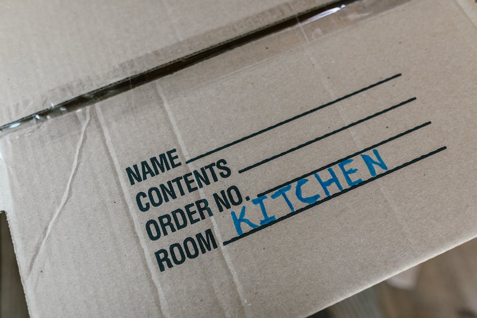 Close-up view of a cardboard moving box labeled with black printed text on a white background, featuring fields for name, contents, order number, and room, with the word 'KITCHEN' handwritten in blue ink in the room section. The box appears to be part of a home relocation process, likely packed for moving services provided by West Hampstead Removals. The background shows additional packing materials and possibly other boxes, indicating an organized packing and loading process inside a property, preparing for a furniture transport or home move from the Fortune Green estate area in NW6. The lighting is natural, showcasing the box in a clear, detailed manner suitable for accessibility and SEO relevant content about packing and moving tips.