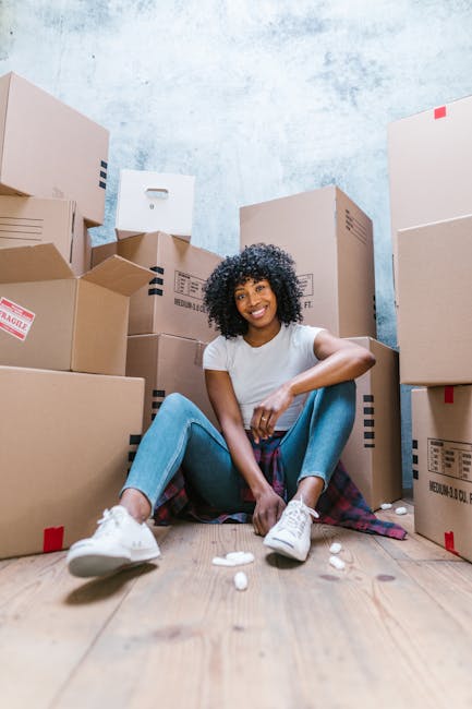 A young woman with curly black hair and dark skin, smiling while sitting cross-legged on a wooden floor surrounded by numerous packed cardboard boxes of various sizes, some with labels and red tape. She is wearing a white t-shirt, light blue jeans, and white sneakers. The boxes are stacked against a textured light blue wall, indicating a home relocation or packing process as part of house removals. In front of her, small packing peanuts and bubble wrap are visible, suggesting careful handling of fragile items. The scene appears to take place indoors, with natural or soft lighting highlighting the organised packing environment. The image reflects the packing stage of a move, related to furniture transport and loading procedures, aligned with services provided by West Hampstead Removals as seen on the NW6 removals tips page for moving from the Fortune Green estate.