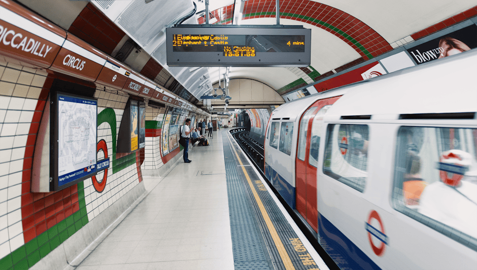 Inside a London Underground station with tiled walls decorated in red, white, green, and black patterns, a packed platform features passengers waiting for a train, with some standing and others sitting on benches. A white and red London Underground train is arriving, with motion blur indicating movement. Overhead, a digital display board shows train arrival times and directions, including a four-minute countdown to Elephant & Castle. To the left, a large map and advertisements are mounted on the tiled wall, and a few luggage and shopping bags are visible among the waiting passengers. The station is well-lit with bright, even lighting, and the platform has safety tactile paving along the edge. Occasionally, the staff or commuters are dressed in casual or work attire, reflecting typical busy underground transport environment. This setting, captured during daytime, supports themes of urban transportation and travel logistics, relevant to home relocation and moving services such as those offered by West Hampstead Removals, which specialise in packing and furniture transport for house moves near West Hampstead station.