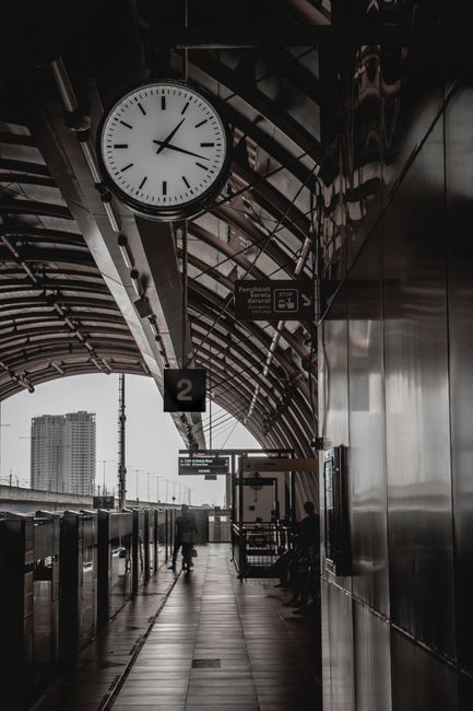 Inside a modern train station platform featuring a large round analog clock displaying approximately 2:11, mounted on a metal support beam attached to a curved, semi-transparent roof structure. The platform is lined with glass barriers and metal railings, with a few passengers visible standing or walking nearby. Several signs are hanging from the roof, including a black square with the number 2 indicating the platform number, and others providing directions and train timings. The station environment is clean, illuminated by natural light filtering through the roof, with the exterior urban landscape visible in the background. This scene captures the typical setting for home relocation or furniture transport logistics, where moving services like those offered by West Hampstead Removals coordinate timing and loading processes around train platforms, assisting clients with efficient packing, loading, and transport of household belongings.