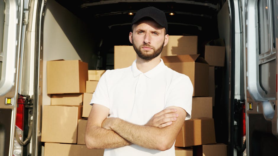 A young man with a beard, wearing a black cap and a white polo shirt, stands inside the open rear of a large moving van. The van is filled with numerous cardboard boxes of various sizes, many sealed with packing tape, stacked neatly to optimise space during a home relocation. The interior of the van is dark, with some boxes positioned on the floor and others stacked on top of each other. Outside, part of a paved area or driveway is visible, suggesting the vehicle is parked outside a property, possibly in West Hampstead. The man has a serious expression, with arms crossed, and is looking directly at the camera. This image relates to furniture transport and packing during a removals service, with West Hampstead Removals providing the logistics for a house move, including loading and organizing boxes for transport. The lighting appears natural, indicating daytime, and the setting is professional and orderly, reflecting typical house removal activities related to managing delays or common problems during same-day moves.