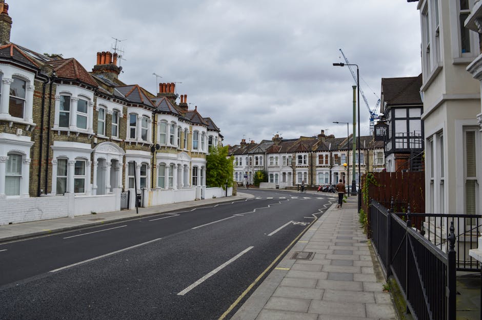 A quiet residential street in West Hampstead showing a row of traditional Victorian-style terraced houses with brick facades, bay windows, and tiled roofs, some with chimneys. The pavement is lined with a black iron fence and a few small plants. The street appears overcast, with grey clouds overhead. In the distance, a moving crane is visible, indicating ongoing construction or renovation work. There are no vehicles or people on the street in the immediate foreground. The photograph depicts an area suitable for home relocation or furniture transport, with clear space for loading and unloading near the houses, and is taken outside during daytime, capturing the typical suburban environment where professional removals, such as those provided by West Hampstead Removals, might operate for house removals or moving services.