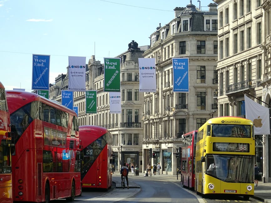 A busy street scene in West End Lane, West Hampstead, featuring three red double-decker buses and a yellow bus parked at a bus stop. The street is lined with multi-storey classic London-style buildings with large windows and ornate facades. Blue and green banners hang across the street, displaying the text 'The West End' and 'London,' with some banners including additional phrases like 'Everyone Welcome.' Pedestrians walk along the pavement, and a cyclist is visible near the buses. In the background, the buildings continue, with some storefronts and residential entrances visible below the banners. An Apple logo flag is also seen, attached to a pole on the right side of the image. The scene is well-lit with natural daylight, capturing the urban environment typical of a central London neighbourhood, suitable for illustrating home relocation or moving services in West Hampstead.